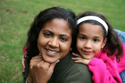 mother with daughter behind with her chin on shoulder, smiling