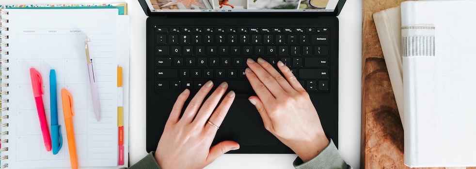 Hands typing on a laptop with a notebook and pen on the desk